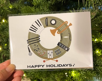 Puede incluir: Una tarjeta ilustrada verde y marrón con un círculo de instrumentos musicales, incluyendo una trompeta, un micrófono, un teclado y una batería. El texto "HAPPY HOLIDAYS" está impreso debajo del círculo.