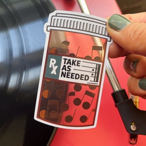 May include: A white sticker with a gold foil design of a pill bottle with the text "Take as Needed" and musical notes. The sticker is on a red surface.