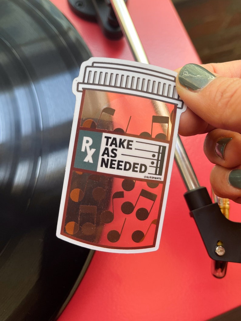 May include: A white sticker with a gold foil design of a pill bottle with the text "Take as Needed" and musical notes. The sticker is on a red surface.