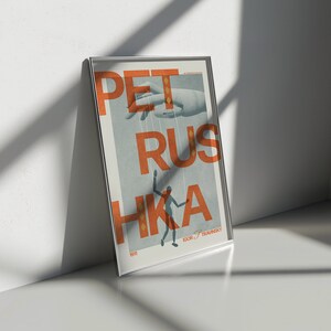 May include: A framed poster with the text "PETRUS HKA" in orange letters on a blue background. A hand with strings attached to it is holding a small figure in the centre of the poster. The text "IGOR "TRAVINSKY" is at the bottom of the poster.