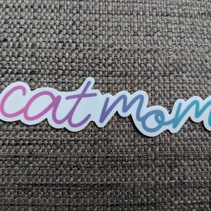 以下が含まれることがあります： 筆記体で「cat mom」と書かれたステッカー。テキストはピンクから紫、そしてティールへと変化します。ステッカーは、質感のある茶色の表面にあります。
