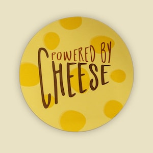 Może przedstawiać: Żółte koło z brązową obwódką i wzorem żółtych kół. Tekst "POWERED BY CHEESE" jest napisany brązowym kolorem na kole.