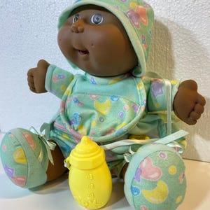 May include: A vintage baby doll with dark skin, dressed in a mint green outfit with a matching bonnet and bib, decorated with pastel shapes. A yellow baby bottle is in front of the doll, and matching booties are on the doll's feet.