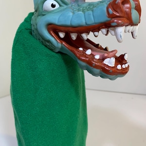 May include: A green hand puppet shaped like a cartoon dragon. The dragon has a blue head with red accents, white teeth, and a wide open mouth. The puppet's body is a solid green color.