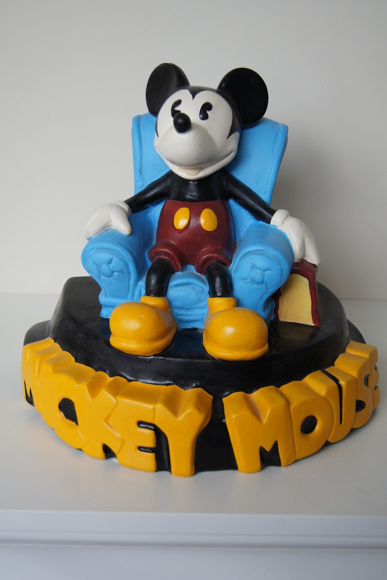 May include: A blue armchair with Mickey Mouse sitting in it. Mickey Mouse is wearing his signature red shorts and yellow shoes. The base of the figurine is black with the words "Mickey Mouse" in yellow letters.