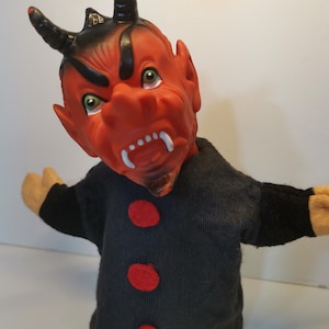 May include: Red devil puppet with black horns and a black shirt with three red buttons. The puppet has a menacing expression with sharp teeth and green eyes.