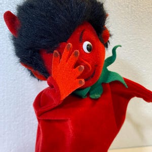 May include: A red devil hand puppet with black hair, red horns, and a green collar. The puppet has a smiling face and is holding its hand up to its face. The puppet is made of a soft, plush material.