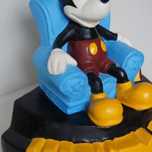 May include: A ceramic figurine of Mickey Mouse sitting in a blue armchair. The figurine is on a black base with the word "Mickey" written in yellow letters.
