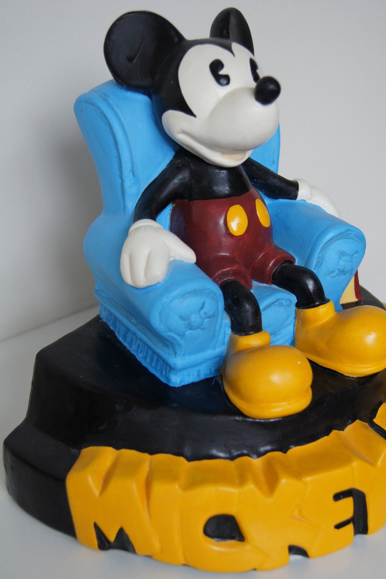 May include: A ceramic figurine of Mickey Mouse sitting in a blue armchair. The figurine is on a black base with the word "Mickey" written in yellow letters.