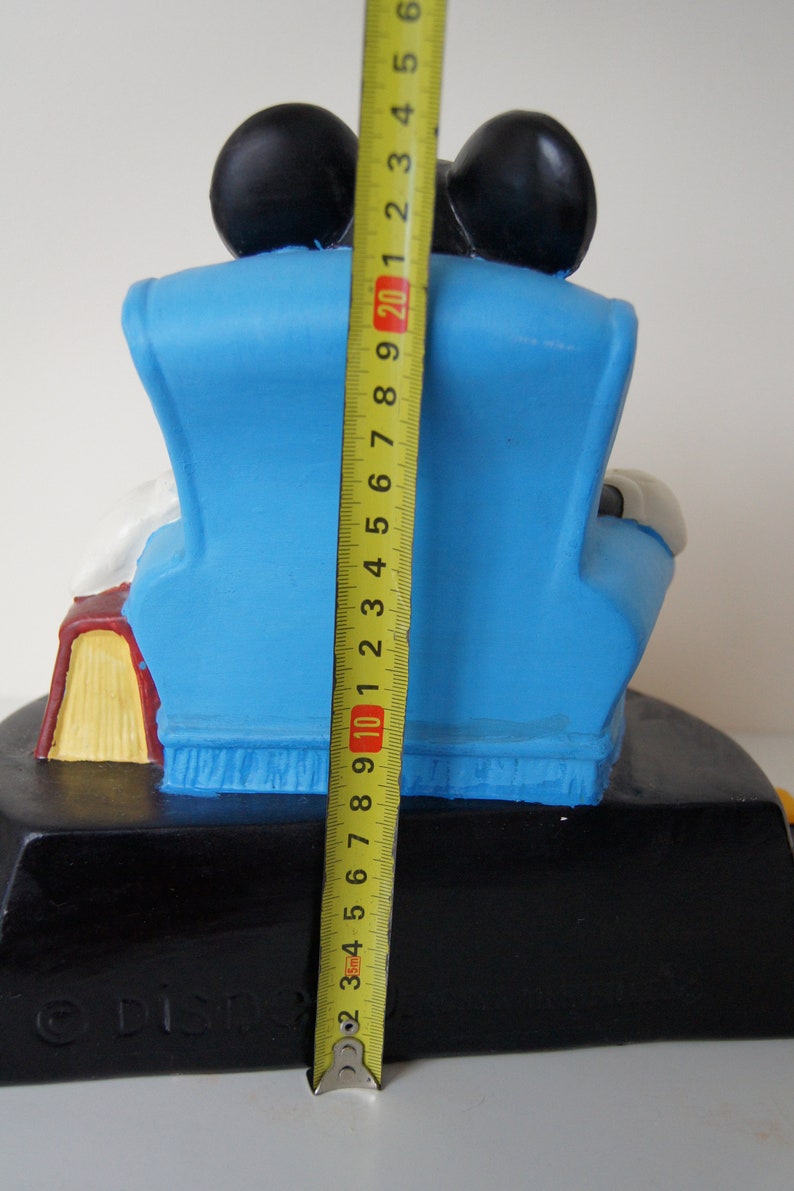 May include: A blue ceramic figurine of Mickey Mouse sitting in a blue armchair with a book in his lap. The figurine is 20 inches tall and is on a black base with the word "DIST" printed on it.