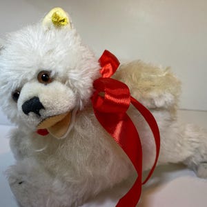 Peut inclure: Un jouet en peluche en forme de chien blanc avec un ruban rouge autour du cou. Le chien a des yeux bruns, un nez noir et des détails jaunes sur les oreilles. Le jouet est fait d'une matière douce et duveteuse.