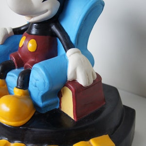 May include: A black and white Mickey Mouse figurine sitting in a blue armchair with a red and yellow book. The base of the figurine is black with the word "MOUSE" in yellow letters.
