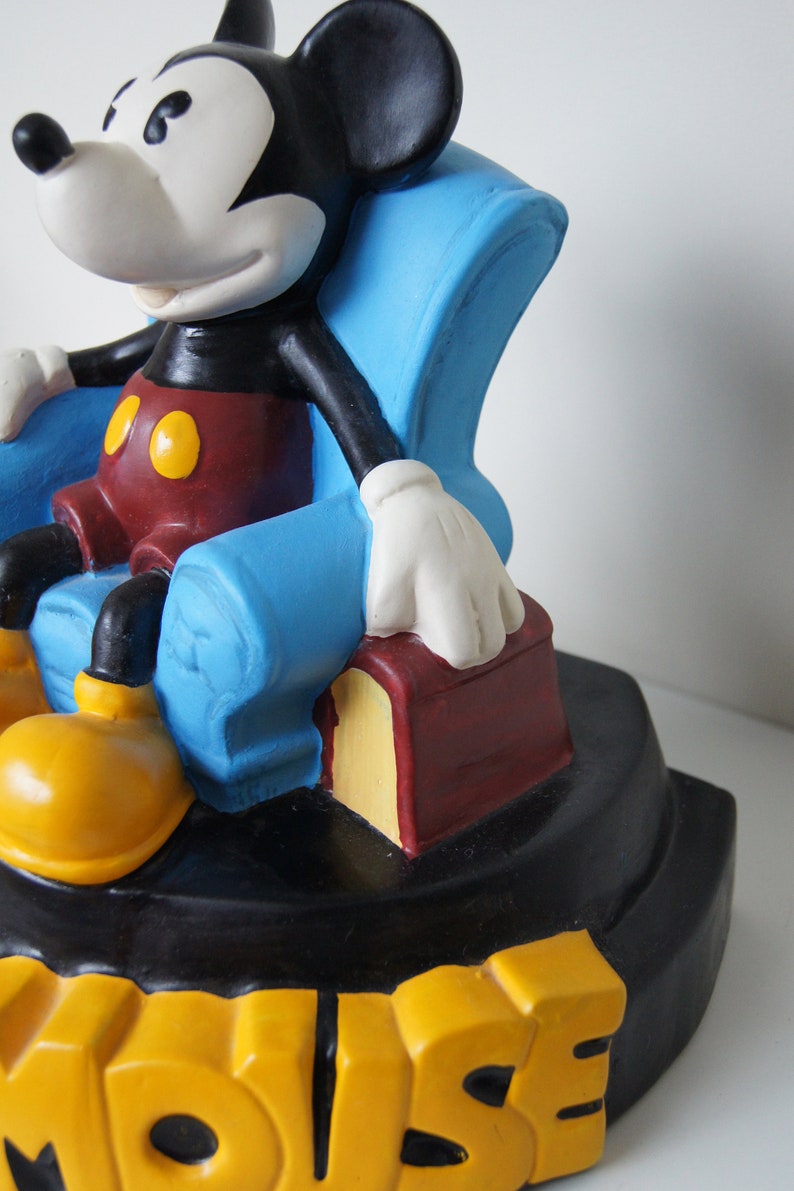 May include: A black and white Mickey Mouse figurine sitting in a blue armchair with a red and yellow book. The base of the figurine is black with the word "MOUSE" in yellow letters.