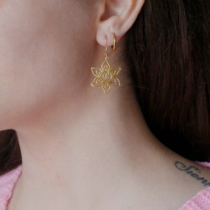 May include: Gold flower-shaped earrings with a small circle in the centre. The earrings are hanging from a person's ear.