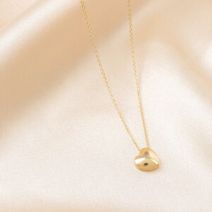 May include: A gold-colored necklace with a heart pendant. The heart is a solid, rounded shape, and the chain is delicate. The necklace is displayed on a cream-colored, silky fabric.
