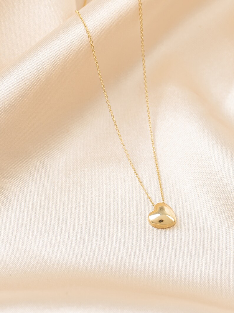 May include: A gold-colored necklace with a heart pendant. The heart is a solid, rounded shape, and the chain is delicate. The necklace is displayed on a cream-colored, silky fabric.
