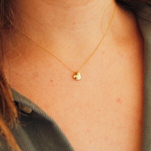 May include: A delicate gold necklace featuring a small, heart-shaped pendant. The pendant hangs from a fine gold chain, resting against a person's skin. The person is wearing a green jacket.