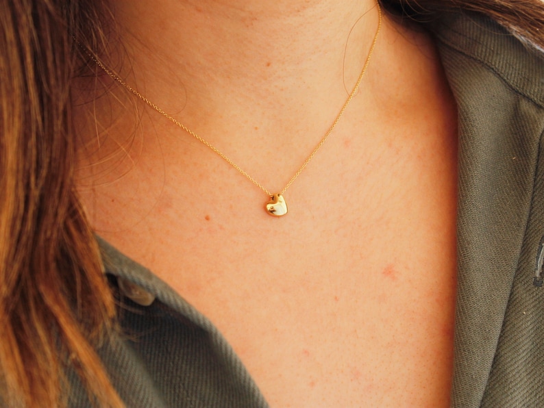 May include: A delicate gold necklace featuring a small, heart-shaped pendant. The pendant hangs from a fine gold chain, resting against a person's skin. The person is wearing a green jacket.