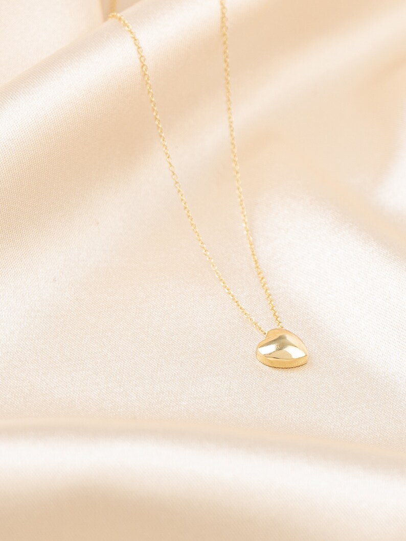 May include: A gold-colored heart pendant necklace. The heart-shaped charm is smooth and rounded, suspended from a delicate chain. The necklace is displayed on a cream-colored satin fabric.