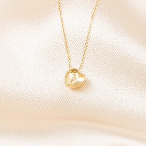 May include: A gold-colored necklace with a heart-shaped pendant. The pendant is small and detailed, with a textured surface. The chain is delicate and thin, and the background is a soft, cream-colored fabric.