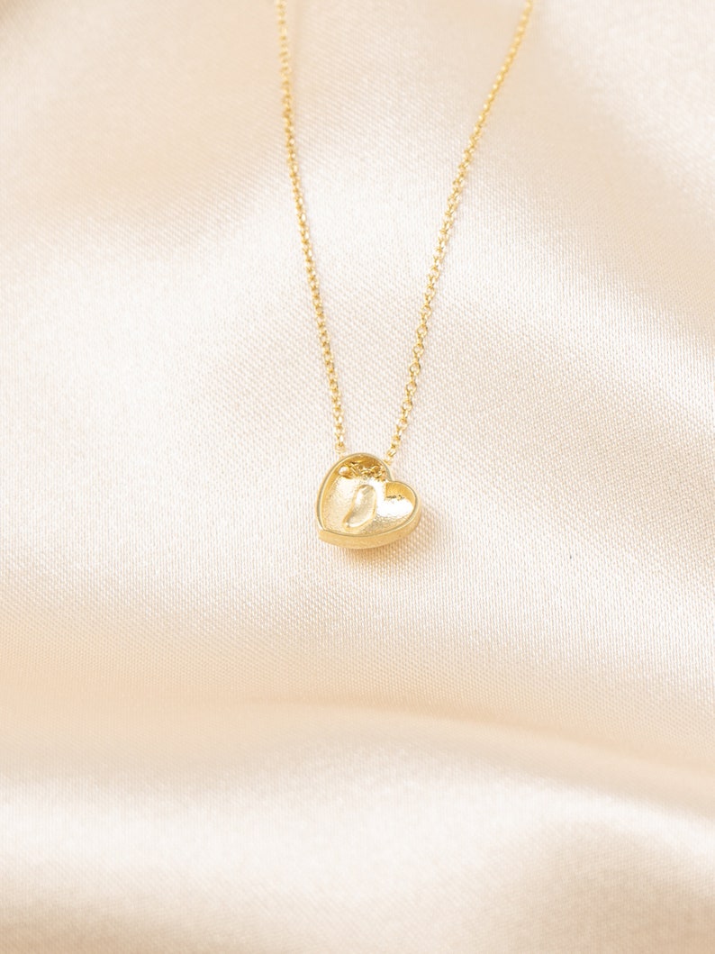 May include: A gold-colored necklace with a heart-shaped pendant. The pendant is small and detailed, with a textured surface. The chain is delicate and thin, and the background is a soft, cream-colored fabric.