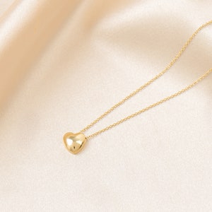 May include: A gold-colored necklace with a heart-shaped pendant. The pendant is smooth and reflective, suspended from a delicate chain. The necklace is displayed on a cream-colored satin background.