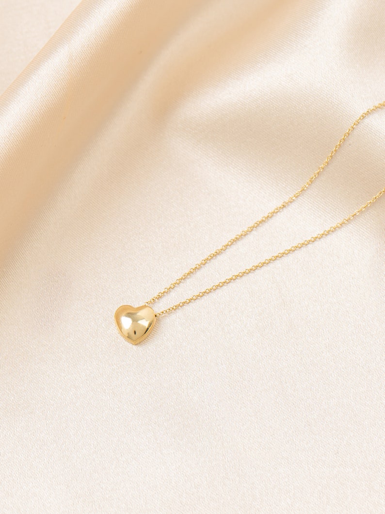 May include: A gold-colored necklace with a heart-shaped pendant. The pendant is smooth and reflective, suspended from a delicate chain. The necklace is displayed on a cream-colored satin background.