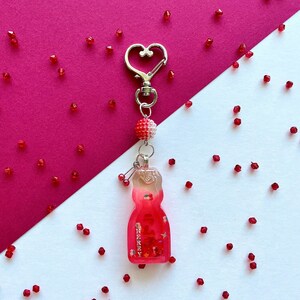 May include: A red and white cherry-flavored gummy bear charm with glitter and a silver heart-shaped keychain. The charm is surrounded by red glitter on a white and pink background.