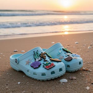 May include: Light blue clogs decorated with beach-themed charms, including a seashell, starfish, and a sign that says "Good the Heat". The clogs are on a sandy beach with the ocean and sunset in the background.
