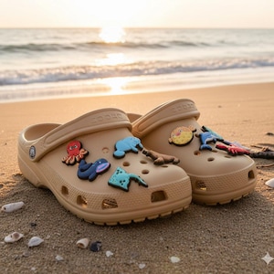 May include: Tan clogs decorated with colorful sea creature charms, including an octopus, whale, and pufferfish. The clogs are on a sandy beach with the ocean in the background. The shoes are a casual style.