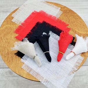 May include: Five square fabric napkins with frayed edges in white, red, and black. Each napkin is folded into a triangle and has a gold ring around it.