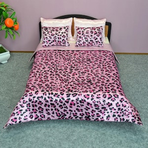 Può includere: Un letto per bambole con testiera nera, con una trapunta rosa con stampa leopardata e federe abbinate. La biancheria da letto ha una finitura satinata. I cuscini hanno federe bianche. Il letto è su un tappeto grigio.