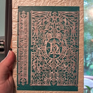 May include: A rectangular book with a textured gold cover and a teal spine panel. The front features an intricate, laser-cut silver overlay with detailed symbols and the text "MAGICAL HIEROGLYPHS & LOGOGRAMS" printed vertically.
