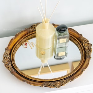May include: A gold oval mirror tray with ornate detailing. The tray has a glass mirror surface and is decorated with a gold-colored frame. The tray is sitting on a white surface and has a diffuser bottle, a perfume bottle, and jewelry on top of it.