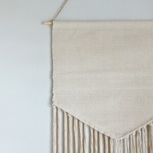 May include: Cream-colored woven wall hanging with a wooden dowel and hanging string. The rectangular textile features horizontal stripes and a triangular bottom edge with long fringe. A simple, neutral home decor item.