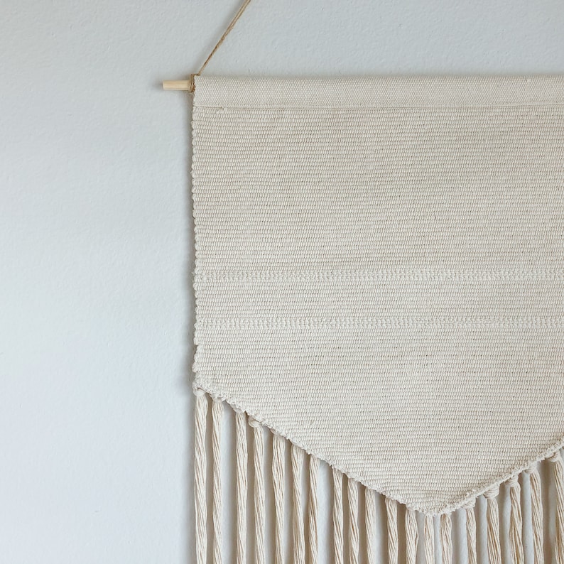 May include: Cream-colored woven wall hanging with a wooden dowel and hanging string. The rectangular textile features horizontal stripes and a triangular bottom edge with long fringe. A simple, neutral home decor item.