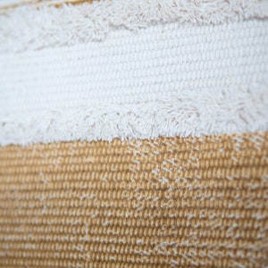 May include: Close-up of a textured fabric with horizontal stripes. The top stripe is off-white with fringe, followed by a white stripe, and a tan stripe at the bottom. The fabric has a woven appearance.