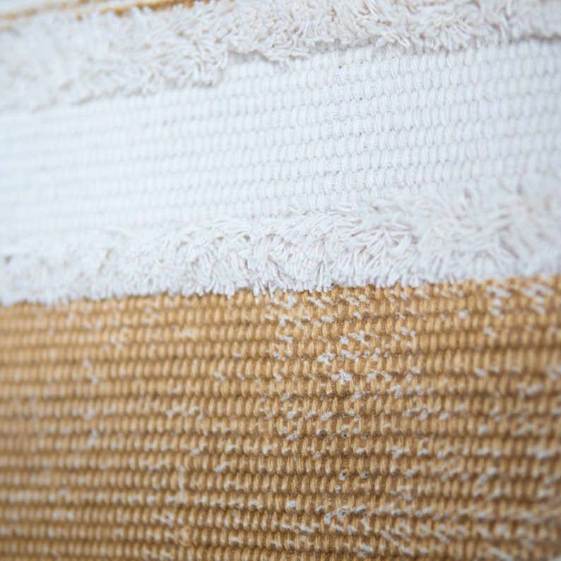 May include: Close-up of a textured fabric with horizontal stripes. The top stripe is off-white with fringe, followed by a white stripe, and a tan stripe at the bottom. The fabric has a woven appearance.
