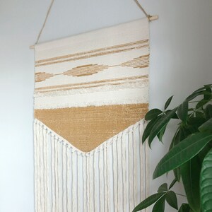 May include: A woven wall hanging with a cream and tan color scheme. The top section features horizontal stripes and diamond patterns in tan. The bottom section is a solid tan color with cream-colored fringe hanging below. The wall hanging is suspended from a wooden dowel.