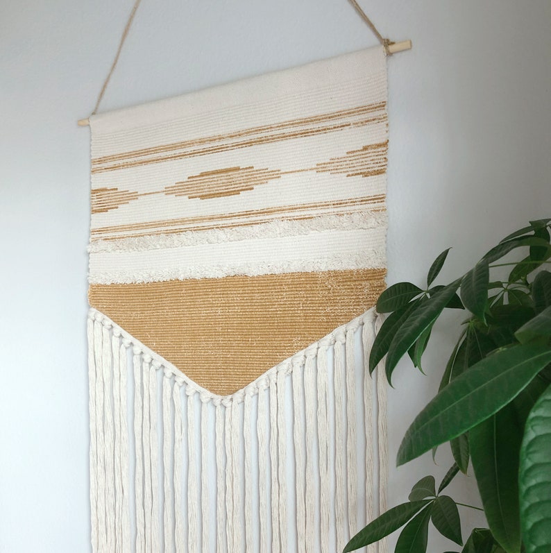 May include: A woven wall hanging with a cream and tan color scheme. The top section features horizontal stripes and diamond patterns in tan. The bottom section is a solid tan color with cream-colored fringe hanging below. The wall hanging is suspended from a wooden dowel.