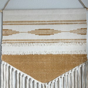 May include: Woven wall hanging with a cream and tan color scheme. The rectangular piece features horizontal stripes, a diamond pattern, and a fringed bottom. It is suspended from a wooden dowel with a natural fiber cord.