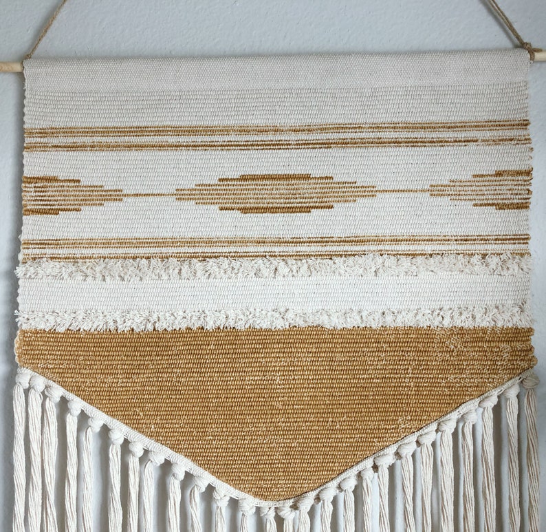 May include: Woven wall hanging with a cream and tan color scheme. The rectangular piece features horizontal stripes, a diamond pattern, and a fringed bottom. It is suspended from a wooden dowel with a natural fiber cord.