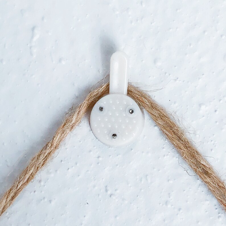 May include: A white picture hanging hook with a textured, circular base and a curved top, designed to hold a picture frame. A length of brown twine is looped through the hook, against a textured white wall.