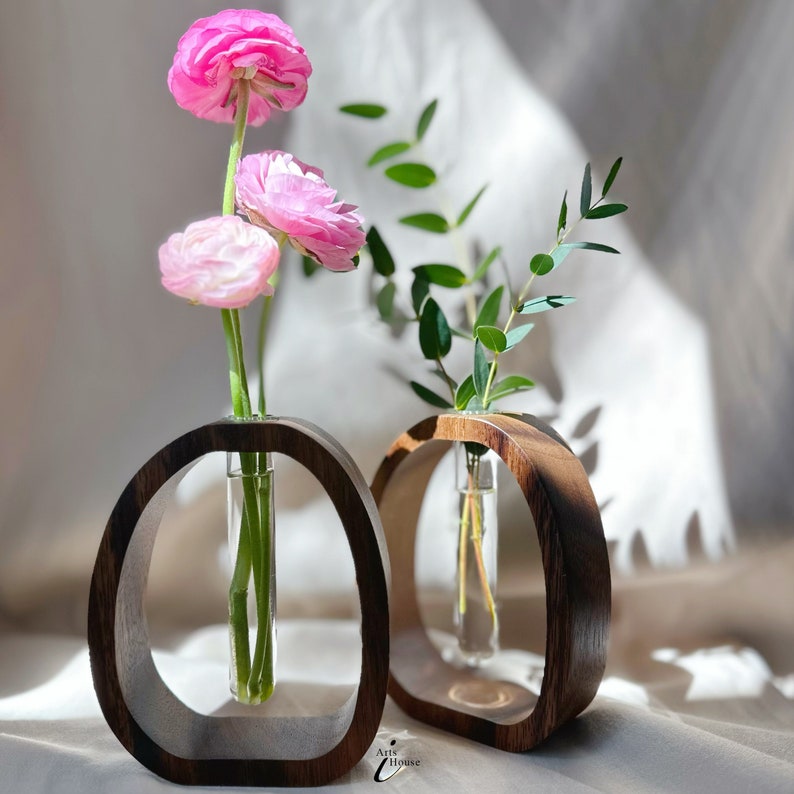 May include: Two wooden vases with glass tubes for holding flowers. The vases are dark brown and have a rounded, oval shape. One vase holds pink flowers and the other holds green leaves.