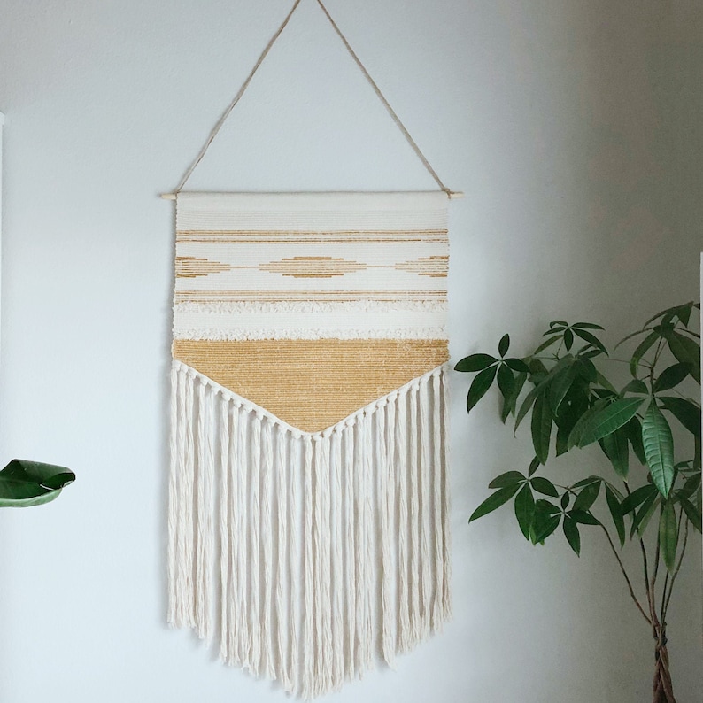 May include: A woven wall hanging with a cream and gold color scheme. The top section features horizontal stripes and diamond patterns, while the bottom has a V-shaped design and long fringe. It is suspended from a wooden dowel by a natural fiber cord.