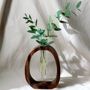 May include: A wooden vase with a round opening, holding a glass test tube with green eucalyptus stems. The vase is brown and has the words "Arts House" inscribed on the bottom.