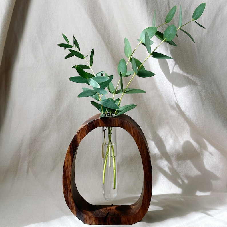 May include: A wooden vase with a round opening, holding a glass test tube with green eucalyptus stems. The vase is brown and has the words "Arts House" inscribed on the bottom.