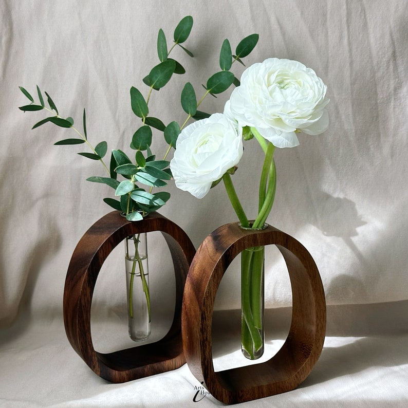 May include: Two wooden vases with a circular opening, each holding a single glass tube with white flowers. The vases are dark brown and have a natural wood grain. The flowers are white ranunculus.