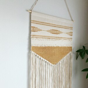 May include: Cream and gold woven wall hanging with a geometric design. The tapestry features horizontal stripes, diamond shapes, and a fringed bottom. It is suspended from a wooden dowel with twine.