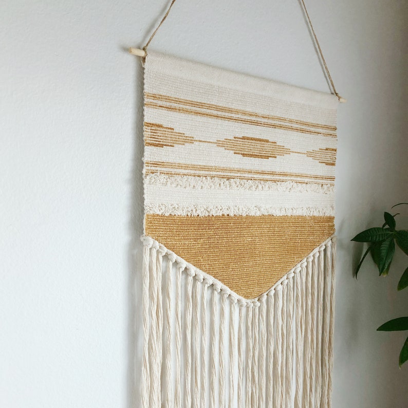 May include: Cream and gold woven wall hanging with a geometric design. The tapestry features horizontal stripes, diamond shapes, and a fringed bottom. It is suspended from a wooden dowel with twine.